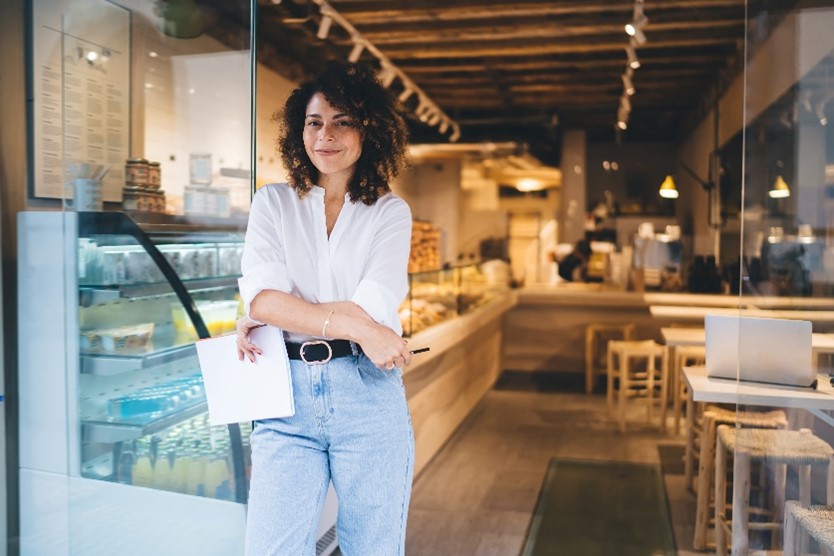 A woman in a white button-down, jeans, and a belt crosses her arms and holds papers while standing at the door of a cafe