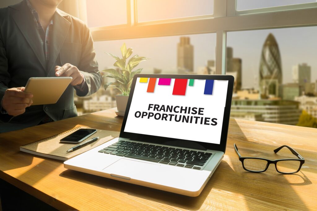 A Laptop on a Table with the Text “Franchise Opportunities” on the Screen and a Person Holding a Tablet in the Background