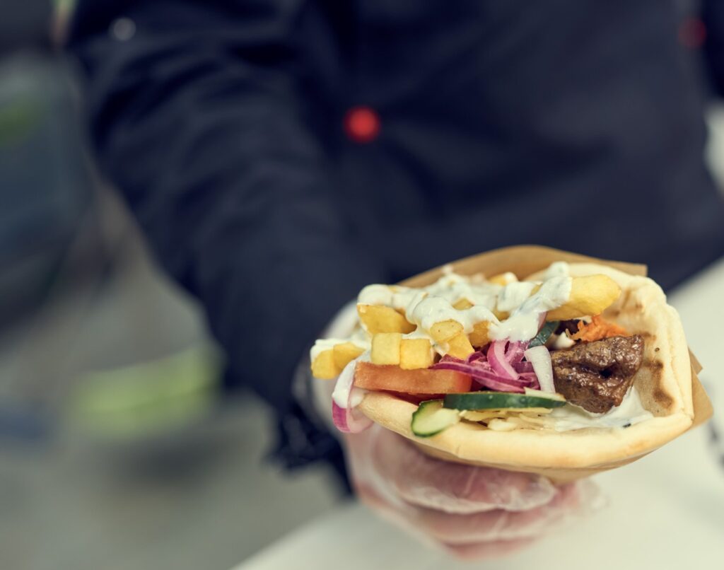 Close Up of a Hand Holding a Pita Sandwich with Various Toppings Including Fries, Steak, Tzatziki, and More