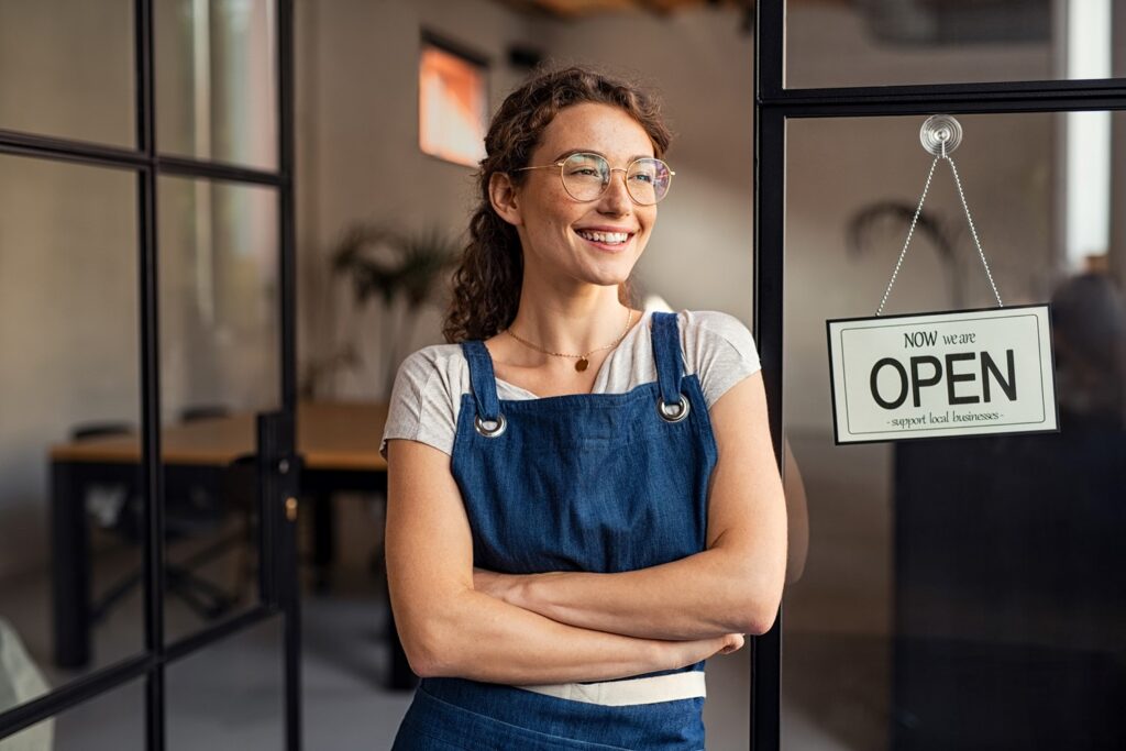 A Smiling Woman Wearing a Denim Apron Stands in Front of an Open Business Door with an Open Sign in the Window Next to Her