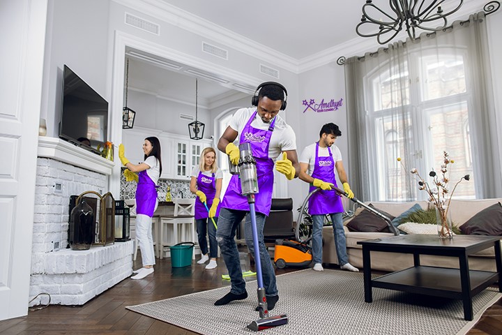 Four cleaners from Amethyst House Cleaning wearing purple aprons while cleaning a living room.