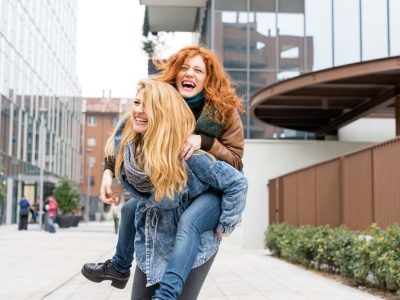 Two young beautiful caucasian blonde and redhead women having fun outdoor in the city riding piggyback - having fun, friendship, carefree concept